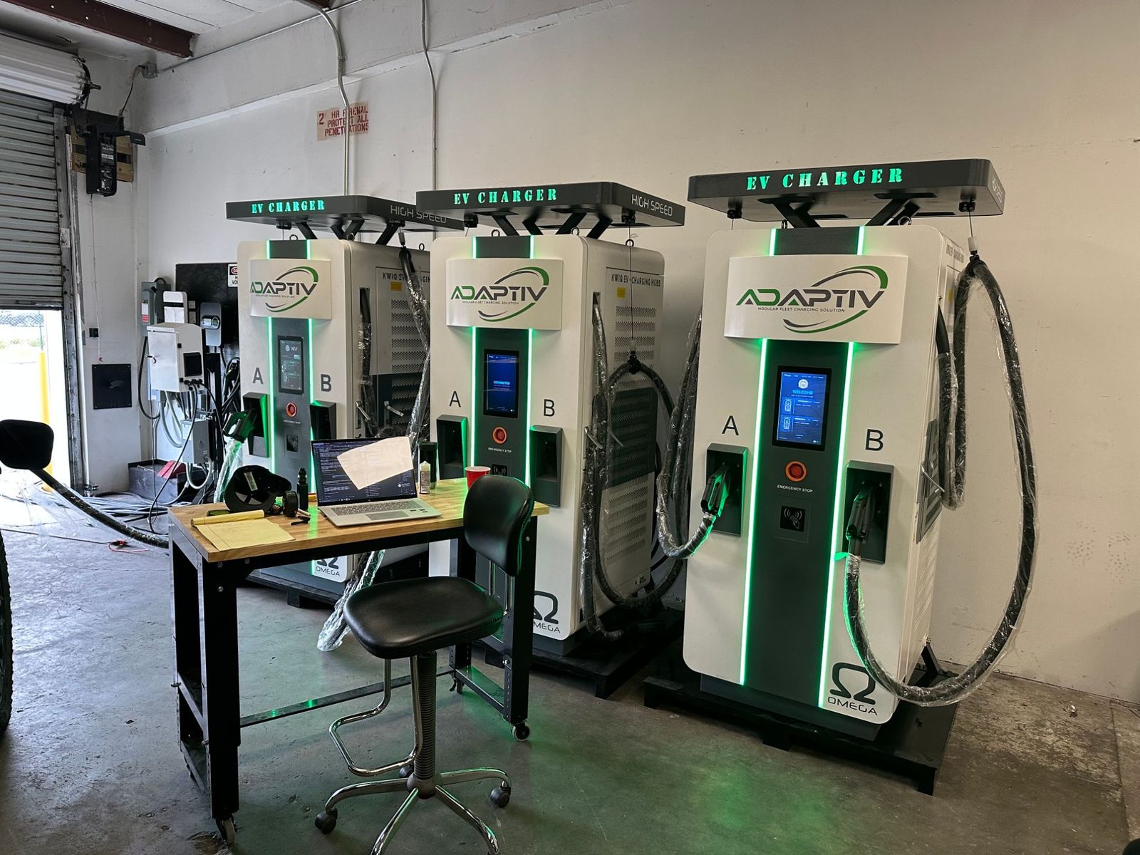 Three dual-port DC fast chargers being commissioned in a workshop with a laptop on a bench connected to the chargers