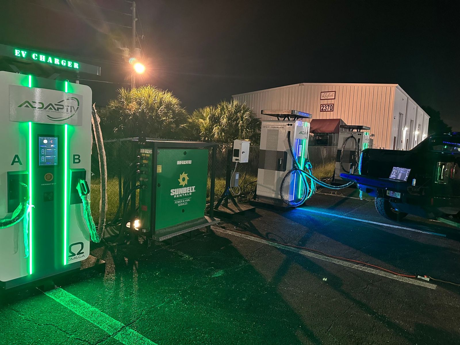 Three dual-port DC fast chargers lit green at night on a temporary pad with a rental power unit behind them