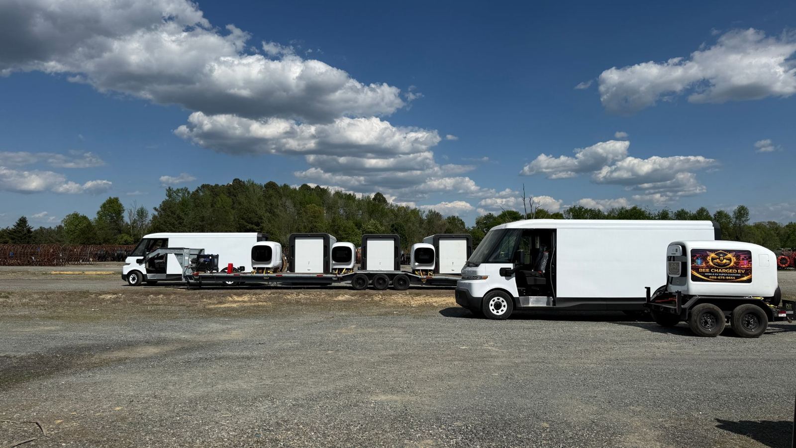 Electric delivery vans and four mobile DC charging pods on flatbed trailers in a gravel yard under a blue sky