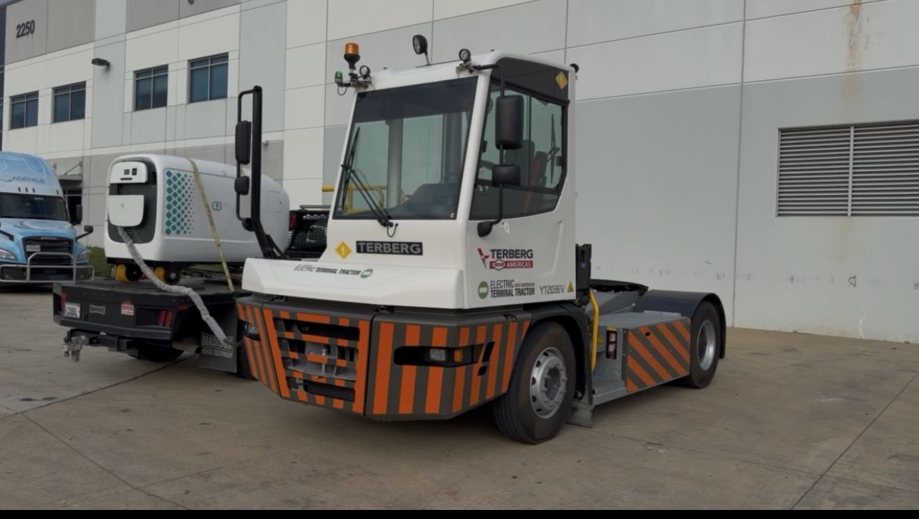 White and orange electric terminal yard tractor plugged into a mobile DC charging pod on a trailer outside a warehouse