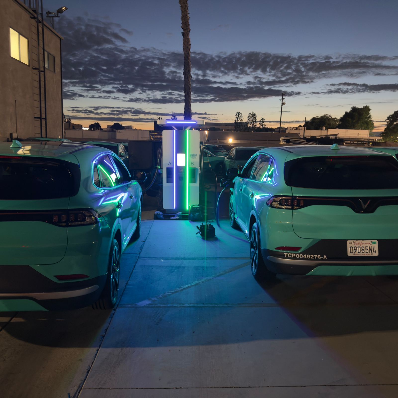 Two teal fleet SUVs at a dual-port DC fast charger lit blue and green in a depot at dusk, more fleet vehicles behind