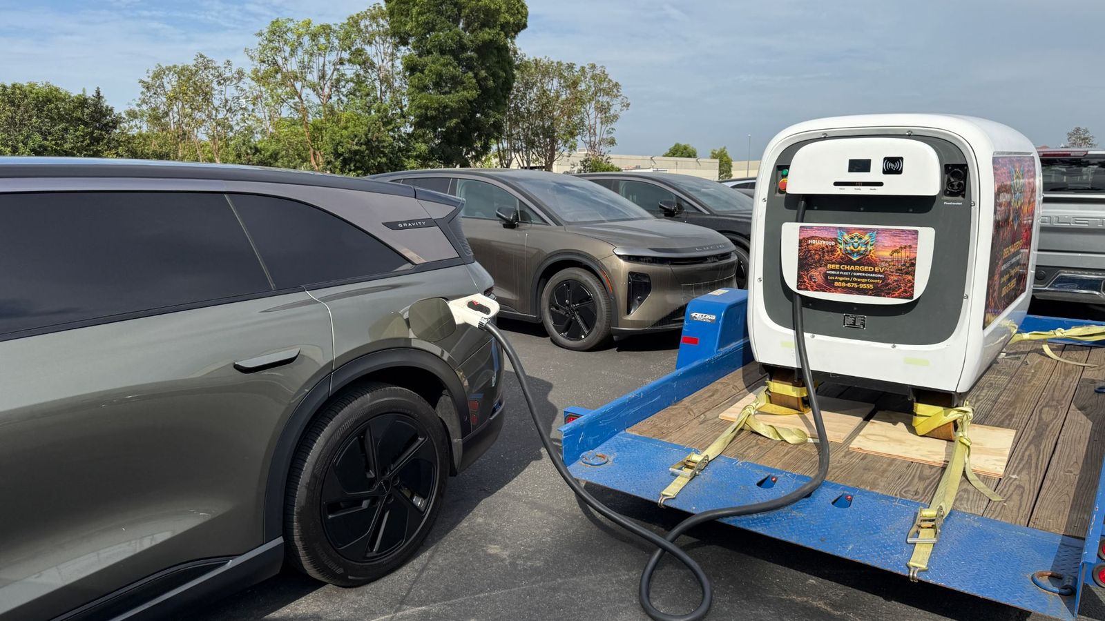 Trailer-mounted mobile DC charger with the CCS cable plugged into a new Lucid Gravity SUV on a vehicle lot, more new vehicles behind