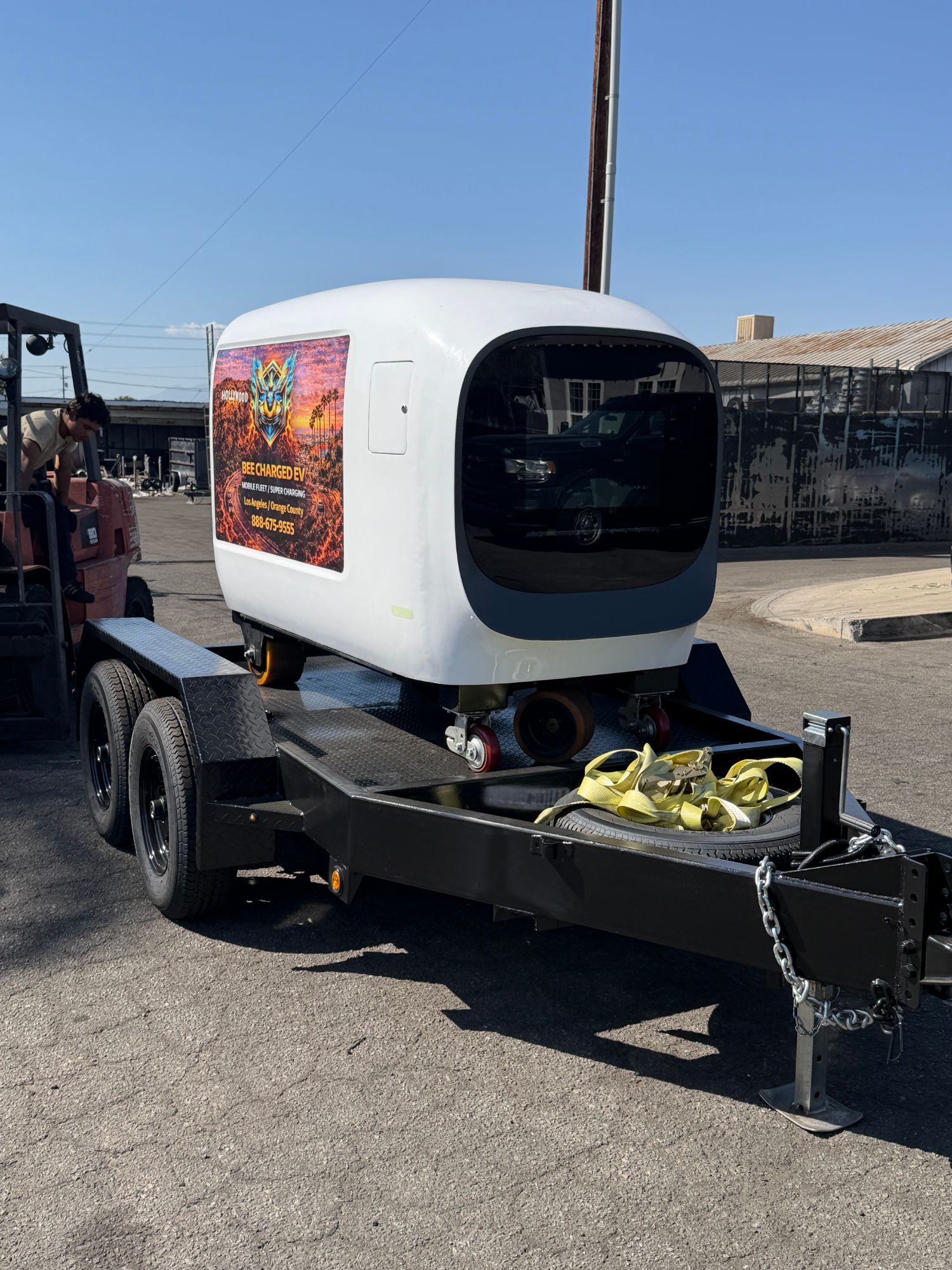 White mobile DC charging pod on casters being loaded onto a tandem-axle trailer with a forklift in a yard