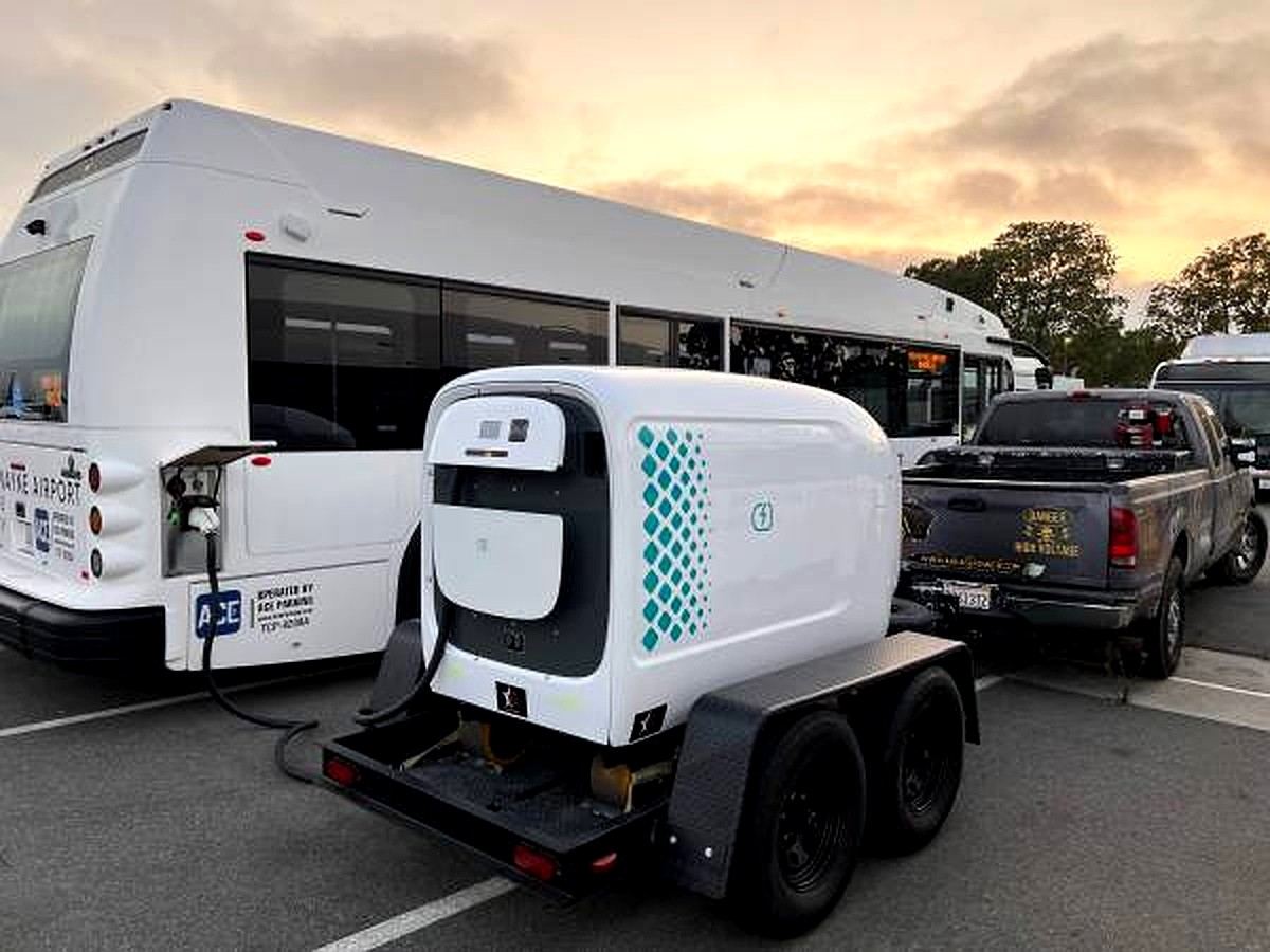 Trailer-mounted mobile DC charging pod plugged into an airport shuttle bus at sunset in a parking lot