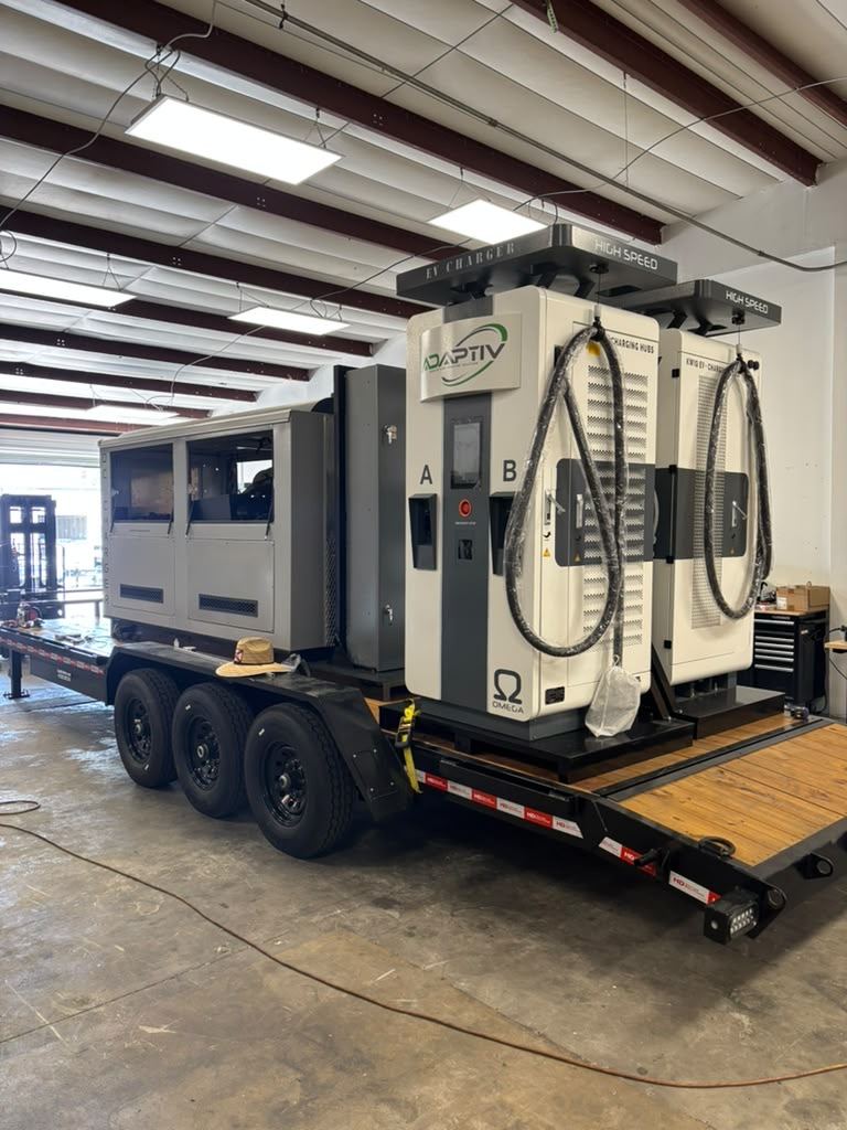 Triple-axle trailer carrying an enclosed power unit and two dual-port DC fast chargers, staged inside a workshop